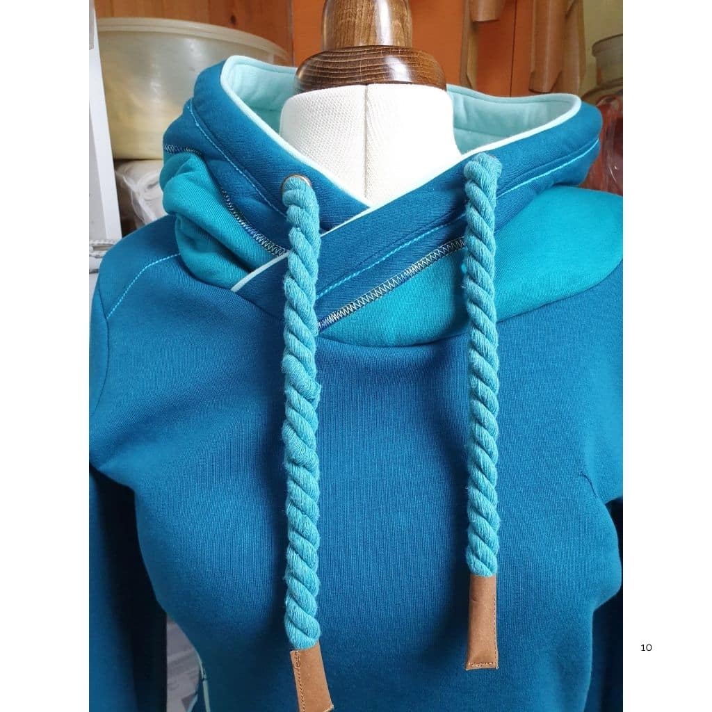 Blauer Hoodie auf einer Schneiderpuppe mit überkreuzter Kapuze, kontrastfarbigem Innenfutter, dekorativen Ziernähten und dicken Kordeln in Türkis.
