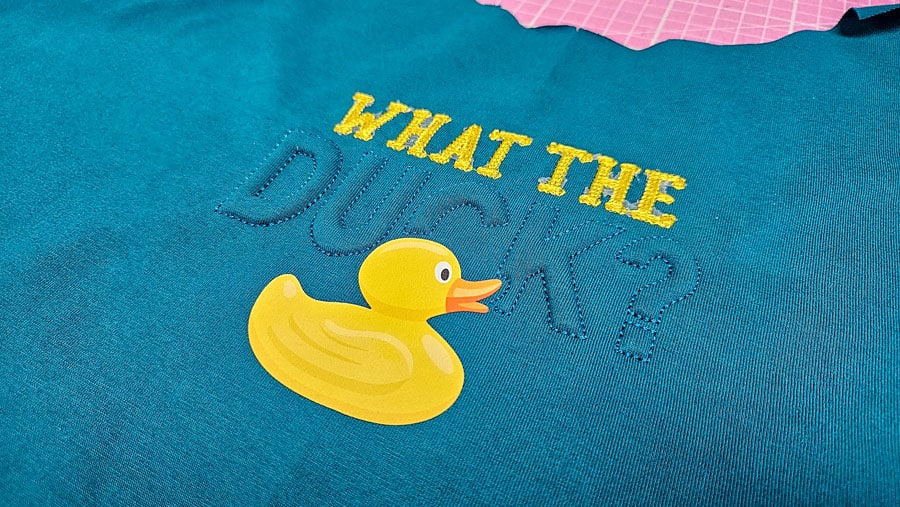 Fertiges Stickmotiv mit gelber Ente, gelber Schrift und plastischer Trapunto-Stickerei auf einem türkisfarbenen Shirt.